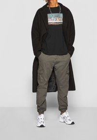 Czarny płaszcz, czarny T-shirt z nadrukiem graficznym, oliwkowe spodnie bojówki, białe i czarne sneakersy, srebrny łańcuszek na szyję.