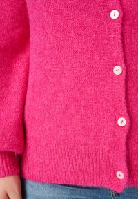Fuchsia gebreid vest met een zachte textuur, voorzien van witte knopen en geribbelde manchetten en zoom, gedeeltelijk weergegeven met daaronder denim stof.