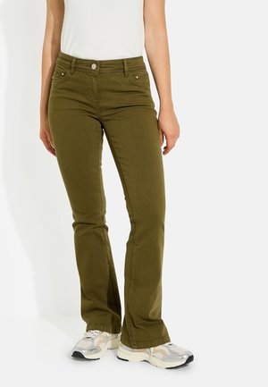 Mujer con pantalones verdes oliva de corte recto y zapatillas blancas, de pie frente a un fondo claro y liso.