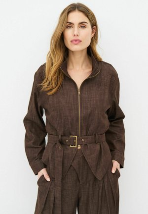 Femme aux longs cheveux portant une veste ceinturée marron foncé et un pantalon assorti, debout les mains dans les poches sur un fond uni.