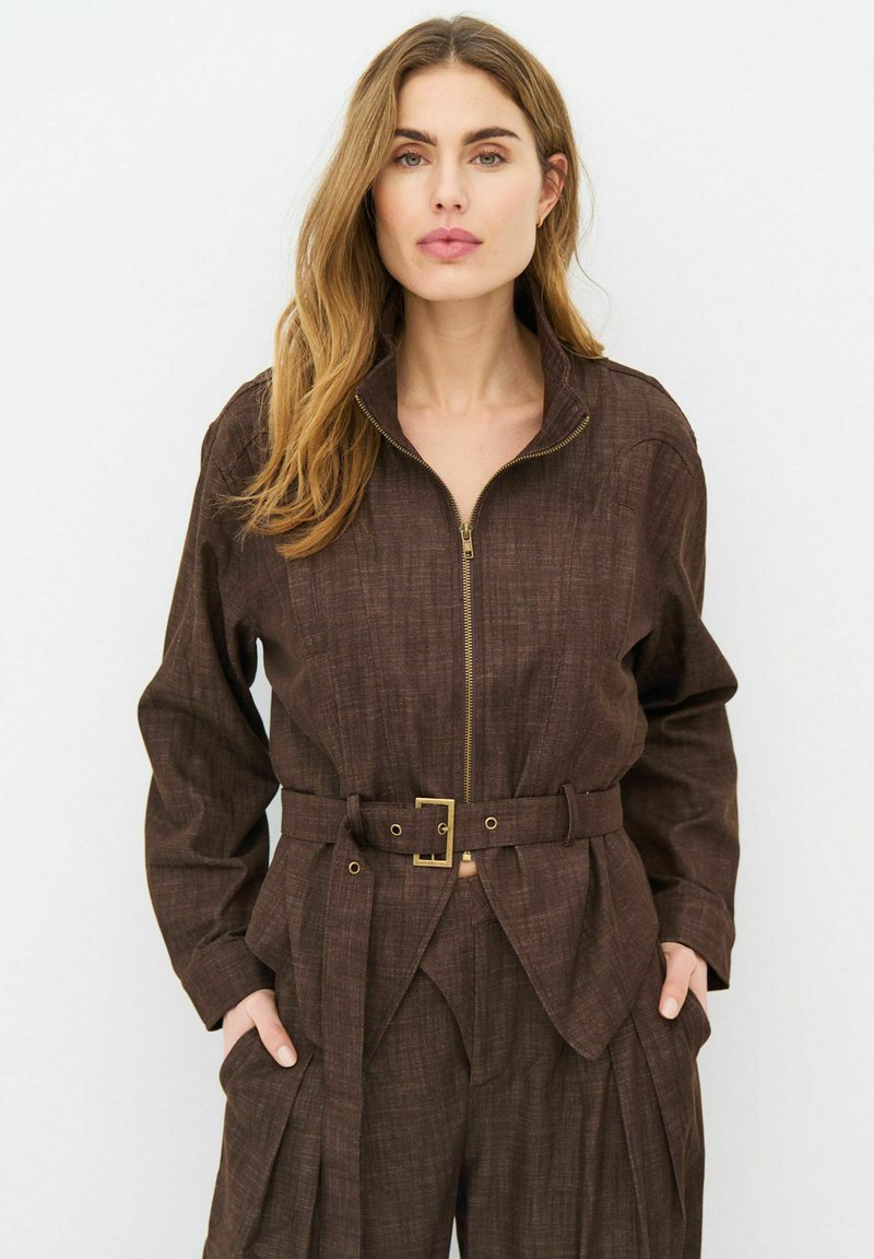 Femme aux longs cheveux portant une veste ceinturée marron foncé et un pantalon assorti, debout les mains dans les poches sur un fond uni.