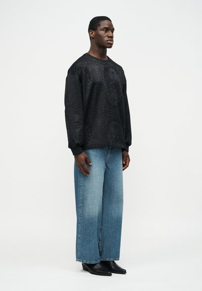 Άνδρας που στέκεται και κοιτάζει προς τα δεξιά, φορώντας μαύρο πουκάμισο με μοτίβο και μακριά μανίκια, φαρδιά μπλε τζιν και μαύρα παπούτσια με μυτερή μύτη.