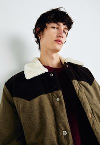 Veste en velours côtelé marron avec un col en fausse fourrure crème, dotée de grands boutons à l'avant et d'un design bicolore, tissu texturé.