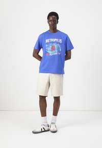Blaues Baumwoll-T-Shirt mit grafischem Design und dem Text "Metropolis" sowie Spider-Man-Motiven, kombiniert mit beigen Shorts und Sneakers.