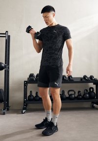 Maglietta atletica nera a motivi, pantaloni corti neri e scarpe da ginnastica nere. Solleva un manubrio. Calze da allenamento grigie con un logo. Attrezzatura da palestra sullo sfondo.