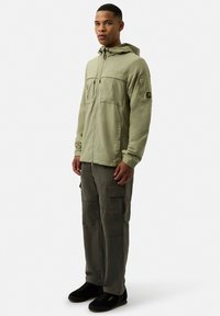 Light green zip-up jacket with a hood and front pockets, paired with dark green cargo pants. Black sneakers with a yellow logo detail.
