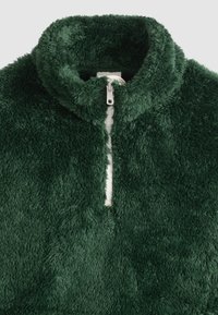 Πράσινο σκούρο φλις μπουφάν με ψηλό γιακά και λευκό φερμουάρ που έχει φερμουάρ μερικώς κλειστό στο κέντρο μπροστά.