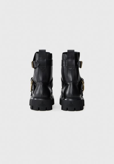 Botines de cuero negro con suela gruesa, acentos de herrajes metálicos en contraste y un detalle de correa en la parte trasera. Textura suave.