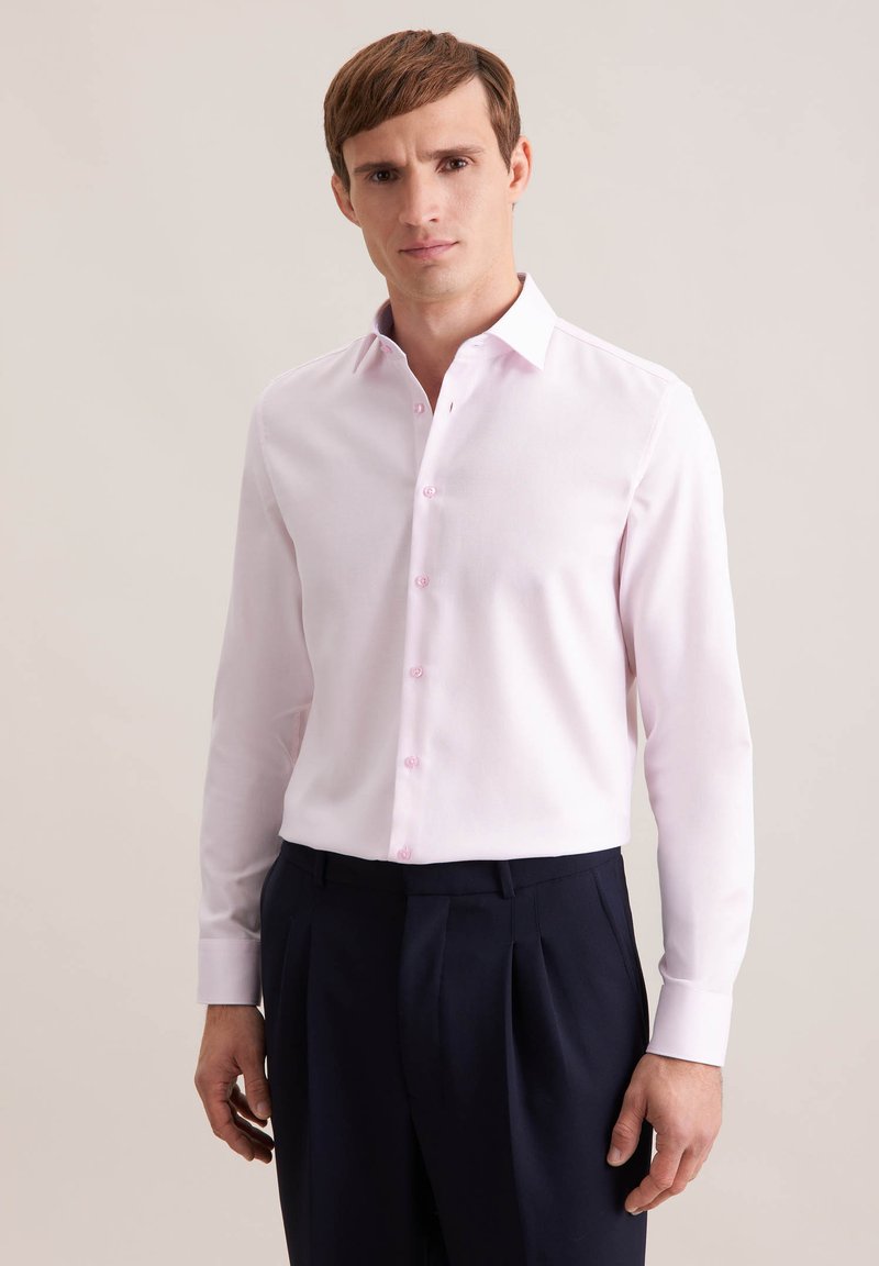 Jeune homme portant une chemise rose clair boutonnée et un pantalon bleu marine foncé, debout devant un fond beige uni.