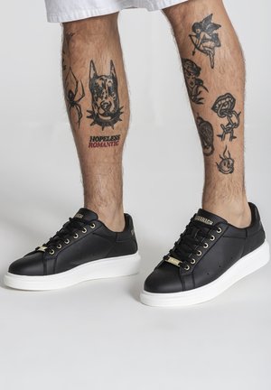 Nohy s čiernymi tetovaniami vrátane hlavy psa, pavúka, cheruba, ruže a textu „Hopeless Romantic“, obuté v čiernych teniskách s bielymi podrážkami.