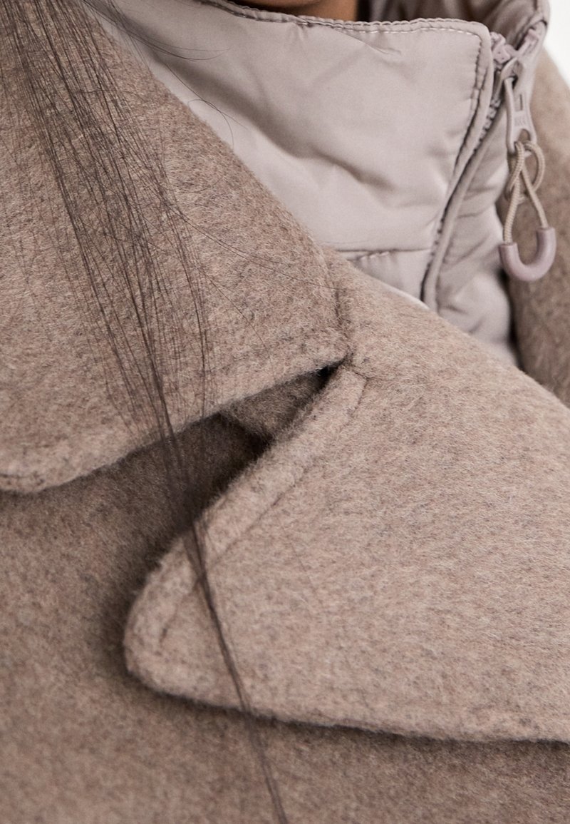 Col de manteau en laine beige superposé sur une veste matelassée beige clair avec une fermeture éclair visible et une languette. De fines mèches de cheveux foncés recouvrent les tissus.