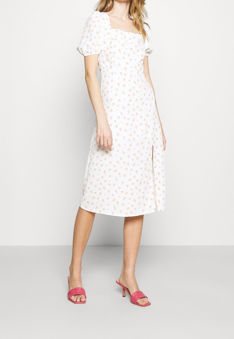 Vestido floral de tela blanca con flores de color naranja claro, escote cuadrado, mangas abullonadas y abertura lateral. Combinado con sandalias de tacón rosas.