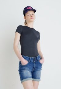 Čierne tričko s krátkym rukávom v kombinácii s modrými riflovými šortkami so zahnutými lemami a prednými vreckami. Model má na hlave čiernu šiltovku s ružovým textom.