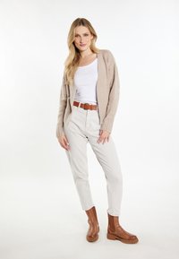 Beige gebreide cardigan met ribbeltextuur over een witte aansluitende tanktop, gecombineerd met lichtbeige broek en bruine enkellaarzen met dikke zolen.