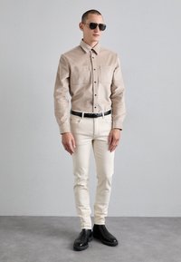 Cămașă bej cu nasturi, cu buzunare aplicate, blugi crem slim-fit și cizme negre până la gleznă. Accesoriile includ ochelari de soare închiși la culoare și o curea neagră.