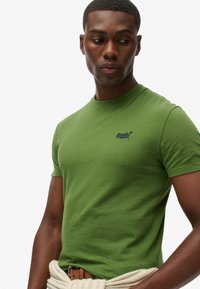 Mand iført grøn Superdry T-shirt, beige sweater bundet om livet, ser nedad og til siden mod en simpel hvid baggrund.