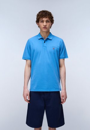 Junger Mann mit lockigem Haar, der ein hellblaues Polohemd und dunkelblaue Shorts trägt, stehend vor einem schlichten hellen Hintergrund.