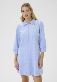 Una donna con capelli ondulati indossa un vestito di velluto a coste azzurro chiaro con colletto e cerniera frontale, in piedi contro uno sfondo bianco semplice.
