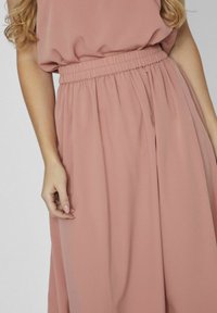 Femme portant un ensemble jupe et haut taille haute de couleur rose poussiéreux avec ceinture élastique, montrant son bras gauche et sa main avec du vernis à ongles clair.