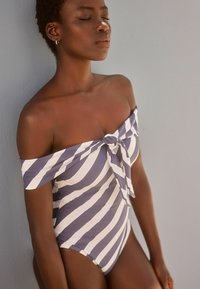 Maillot de bain asymétrique à rayures bleu marine et blanches, avec un détail noué à l'avant et un tissu doux et extensible. Texture lisse, design ajusté.