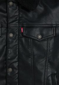 Black leather jacket with a textured finish, featuring a shearling collar, metal snap buttons, and a small red label on the left side.