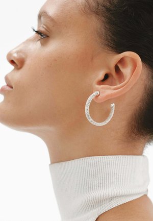 Perfil de una mujer que lleva un pendiente de aro plateado texturizado y un suéter de cuello alto blanco de canalé sobre un fondo liso.