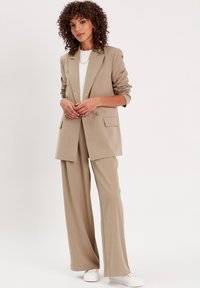 Blazer beige su misura e pantaloni a gamba larga, con tessuto morbido, due tasche frontali e vestibilità rilassata, abbinati a sneakers bianche.
