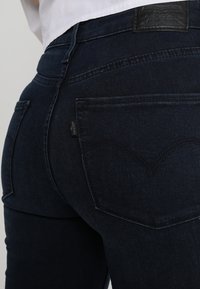 Jeans de color azul oscuro con cintura alta, que cuentan con cinco bolsillos, un parche de marca de cuero y costuras curvadas en los bolsillos traseros.
