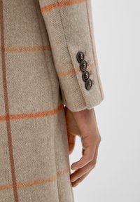 Abrigo de lana beige con un patrón de cuadros naranja, superficie texturizada y cuatro botones oscuros en el puño. Mano colocada en la manga.