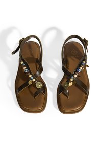 Braune Ledersandalen mit Kreuzriemendesign, verziert mit dekorativen Perlen und Charms entlang des Riemens sowie einer strukturierten braunen Sohle.