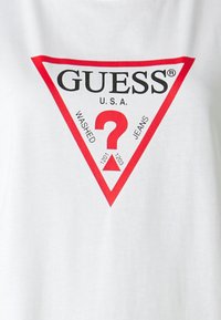 Bílý bavlněný tričko s velkým červeným trojúhelníkovým logem a černým textem, který zní "GUESS U.S.A. WASHED JEANS" a otazníkem.