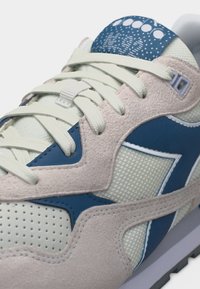 Κοντινό πλάνο αθλητικού παπουτσιού με κορδόνια κρεμ, επενδύσεις από μπεζ σουέντ, πάνελ από κρεμ δίχτυ, και μπλε δερμάτινες λεπτομέρειες, που δείχνει την λεπτομερή υφή και τη ραφή.