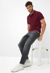 Polo shirt bordeaux con maniche corte, abbinato a jeans grigi scuri e sneaker bianche. Le calzature presentano accenti beige e grigi.