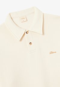 Kermanvärinen polo-paita, jossa on pehmeä rakenne, kaulus, kaksi nappia ja pieni kultainen brodeerattu logo.
