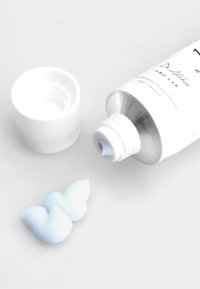 Hvit kremtube med sølvhette, delvis åpen, med lys blå krem som er presset ut i en spiralform på en lys grå overflate.