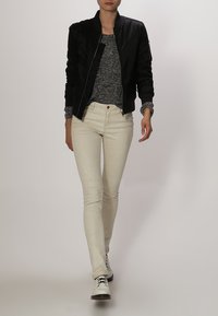 Giubbotto bomber nero con zip frontale, maglione grigio a maglia, pantaloni skinny bianchi con un motivo a macchie e scarpe bianche con lacci neri.