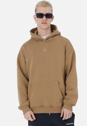 Brown hooded sweatshirt made of soft material, featuring a front pocket and ribbed cuffs, worn with a white shirt and sunglasses.