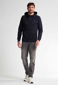 Felpa con cappuccio blu navy con zip e cordoncino, indossata sopra una maglia nera. Abbinata a jeans grigi e sneakers bianche per un look casual.