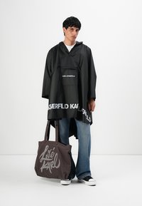 Mlad čovjek nosi crnu ponču Karl Lagerfeld s bijelim logotipom, plave traperice, crno-bijele tenisice i drži veliku tamnu torbu.