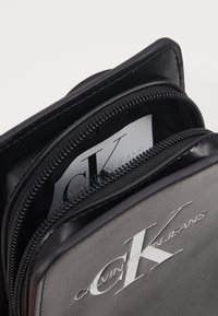 Černá taška Calvin Klein Jeans s dvěma zipovými přihrádkami, jedna částečně otevřená, ukazující značkovou etiketu uvnitř.