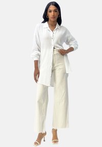 Chemise blanche texturée à boutons avec des manches longues, associée à un pantalon blanc à taille haute et à jambes larges, et des sandales à talons nus.