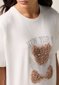 Witte katoenen T-shirt met een fuzzy bruine beer graphic, versierd met rhinestone accenten en de tekst "NEW YORK" in kristalversieringen.