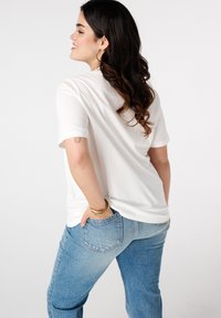 Witte t-shirt met korte mouwen, ontspannen pasvorm, gecombineerd met lichtblauwe high-waisted jeans. Zichtbare gouden armbanden om de pols. Eenvoudig ontwerp, gladde textuur.