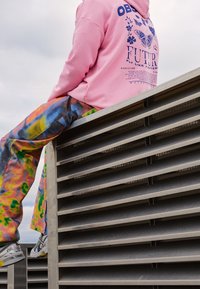 Sudadera rosa con un diseño gráfico y texto en la parte de atrás, combinada con pantalones de tie-dye coloridos. Sentado sobre una estructura metálica con lamas.