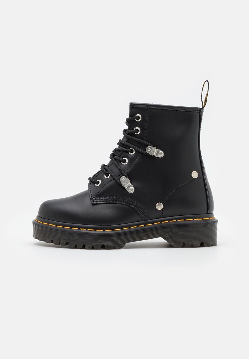 Botte de combat en cuir noir avec semelle épaisse, coutures jaunes, œillets métalliques et languette à l'arrière, vue de côté.