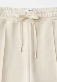 Light beige sweatpants with an elastic waistband, drawstring detail, and a subtle texture. Features a logo tag at the waist.