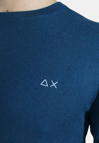 Maglione blu con una texture a coste e un collo rotondo. Piccolo logo geometrico bianco sul lato sinistro del petto. Tessuto morbido.