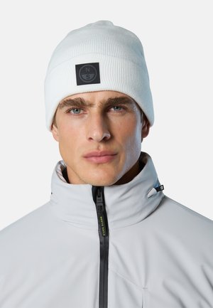 Giovane uomo con occhi azzurri che indossa un berretto lavorato a maglia bianco con un logo nero e una giacca bianca con collo alto chiusa con cerniera.