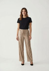 Mujer de pie y sonriendo con cabello ondulado, lleva una camiseta negra de manga corta, pantalones beige con estampado de leopardo y tacones negros sobre un fondo liso.