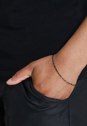 Bracciale a catena nera con design intrecciato indossato al polso, la mano poggia in tasca, mettendo in risalto la sua delicata texture e il sottile luccichio.